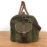 A White Cap Waxed Canvas Duffle from Rogue Industries in green, featuring brown leather accents, a front pocket with a buckle closure, and a full-grain leather shoulder strap, sits on a wooden surface against a plain white background. The water-resistant waxed canvas ensures durability and style.