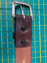 Close-up of a handcrafted Baxter Belt and Made in Maine Rogue Wallet Bundle by Rogue Industries with a silver buckle, featuring black screws securing the buckle. A green cutting mat with yellow grid lines serves as the background.