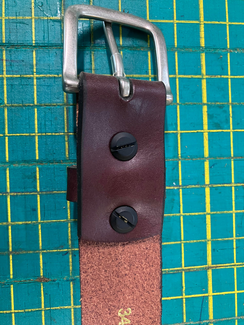 A close-up image of a leather belt with a metal buckle, featuring two black screws securing the leather end. The background shows a cutting mat with a green grid pattern. This Chamberlain Belt and Minimalist Wallet Bundle from Rogue Industries is crafted with precision, perfect for those who appreciate fine details in their accessories.