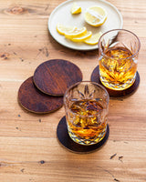 Two glasses of whiskey on a wooden table, resting on a Rogue Industries Moose Leather Coaster Set.