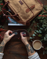 A person holding a Rogue Industries Waxed Canvas Laptop Bag next to a durable wallet and a cup of coffee.