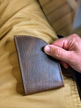 A hand is holding a slim Factory Second PREVIOUSLY EMBOSSED Made in Maine Front Pocket Wallet - BROWN by Rogue Industries against a background of khaki-colored pants. This stylish accessory, crafted in Maine, features RFID-blocking technology for added security.