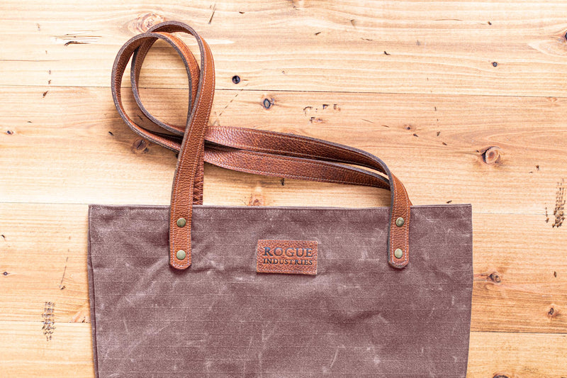 A durable, brown waxed canvas Allagash River Tote Bag on a wooden floor by Rogue Industries.