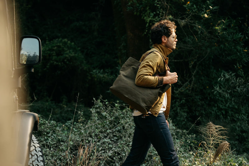 A man walking in the woods, carrying a durable Rogue Industries Waxed Canvas Tote Bag with top-grain leather straps.