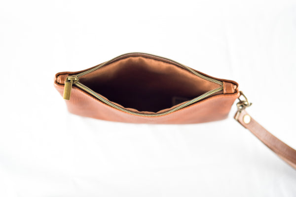 Top view of an open Sebago Leather Clutch by Rogue Industries, crafted from top-grain brown leather, featuring a prominent metal zipper and a detachable strap, set on a white surface. Made in the USA.