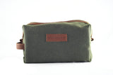 A green fabric toiletry bag with brown leather accents, crafted as a Waxed Canvas Dopp Kit, and featuring the Rogue Industries label on the front. Made in the USA.