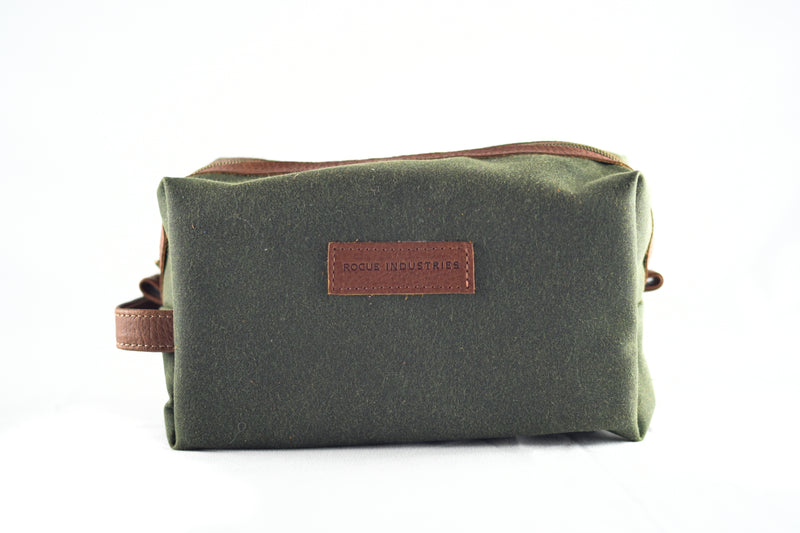 A green fabric toiletry bag with brown leather accents, crafted as a Waxed Canvas Dopp Kit, and featuring the Rogue Industries label on the front. Made in the USA.