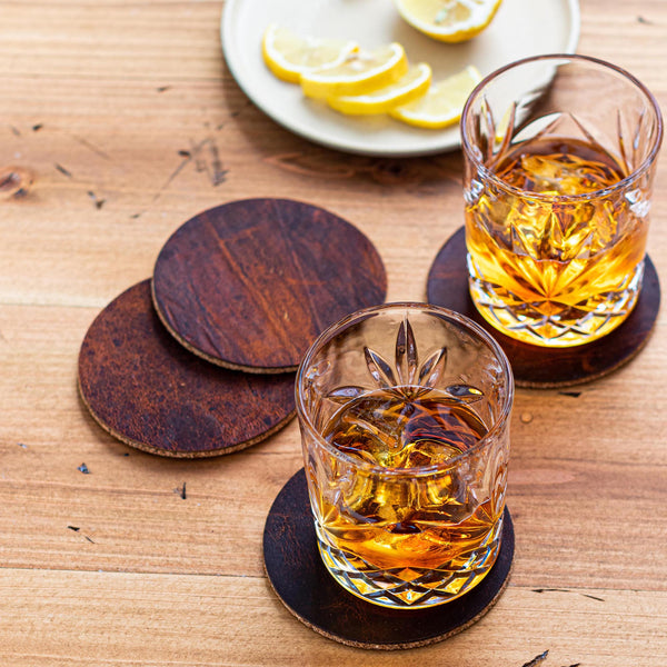 Two glasses of whiskey on a wooden table, resting on Rogue Industries Genuine Moose Leather Coaster Set.