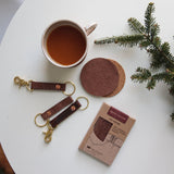 A cup of coffee, Rogue Industries' Salmon Leather Coaster Set, a genuine salmon leather keychains, and a packaging box on a table, suggesting a styled product display with a cozy atmosphere.